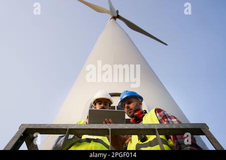 Two helmeted engineers using and looking tablet. Professional technical workers at wind power plant. Stock Photo