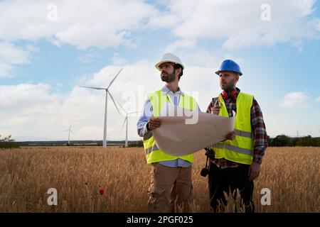 Two engineers in hard hats work holding plane in agricultural field with wind turbines. Outdoors. Stock Photo