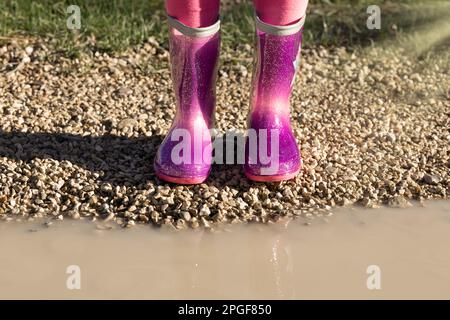 Child wearing new bright pink rain rubber boots standing near muddy puddle, no face. Preschool in wellies. Footwear for rainy weather Stock Photo