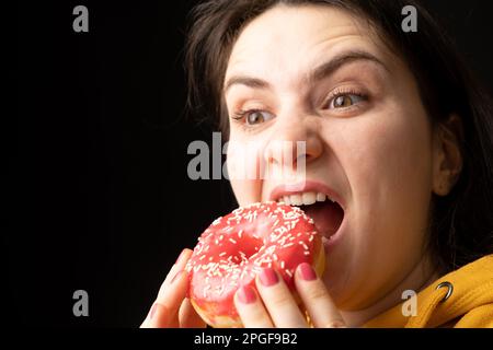 A woman bites a large red donut, a black background, a place for text. Gluttony, overeating and sugar addict. Stock Photo