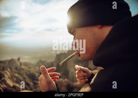 Boy smoking a marijuana joint on top of a mountain during sunset Stock Photo