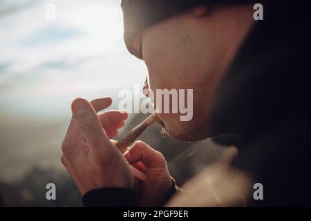 Boy smoking a marijuana joint on top of a mountain during sunset Stock Photo