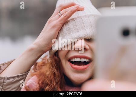 Curly redhead woman 30-35 with a hat over her eyes take selfie Stock Photo