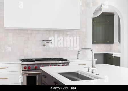 A kitchen detail with a sink on an island's marble countertop with a stove, hood, and tiled backsplash in the background. Stock Photo