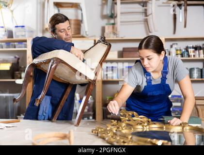 Male and female furniture makers diligently working on vintage mirror and chair in workshop Stock Photo