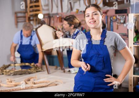 Portrait of smiling skilled young European female furniture repair specialist in uniform posing in workshop Stock Photo