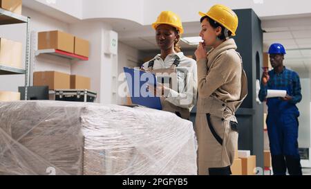 Multiethnic team of women organizing products in warehouse, looking at packages on shelves and counting merchandise stock. People checking stock of goods to prepare shipment order. Stock Photo