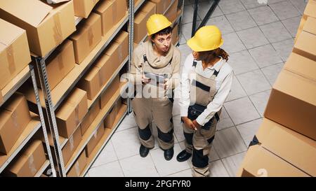 Two women in overalls working with industrial goods, planning shipment of products with digital tablet. Team of employees doing inventory in storage room, manufacturing products. Stock Photo