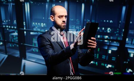 Datacenter engineer looking at mainframe diagnostics using digital tablet, inspecting cloud computing server room. IT infrastructure specialist working with fintech in data center. Stock Photo