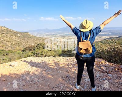 Adult woman with hat looks towards the horizon on top of a mountain opens his arms and breathes in front of the mountainous landscape proud Stock Photo