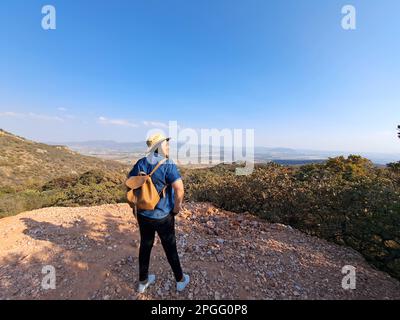 Adult woman with hat looks towards the horizon on top of a mountain opens his arms and breathes in front of the mountainous landscape proud Stock Photo