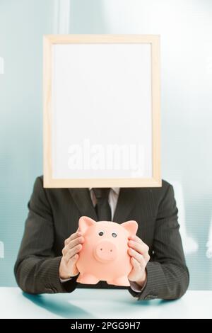 Office workplace, a sign-headed man framed in light wood holds a piggy bank. He holds it carefully, for a concept of savings, spending, investment and Stock Photo