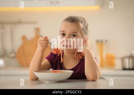 Cute little girl eating tasty pasta at table in kitchen Stock Photo