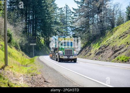 Big rig classic green semi truck tractor with oversize load sign on the roof transporting oversized equipment on step down semi trailer driving on the Stock Photo