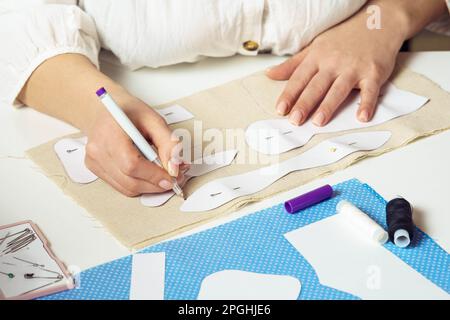 Cropped female seamstress drawing contours with pen of pinned paper sewing patterns on textile sheet on table. Needlework, making patterns for future Stock Photo