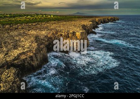 The cliffs on which the stormy waves of the sea break along the coast of the Plemmirio Nature Reserve. In the background, the Etna volcano. Sicily Stock Photo