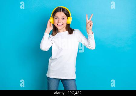 Photo of cheerful friendly positive girl wear white long sleeve showing v-sign touching headphones isolated on blue color background Stock Photo