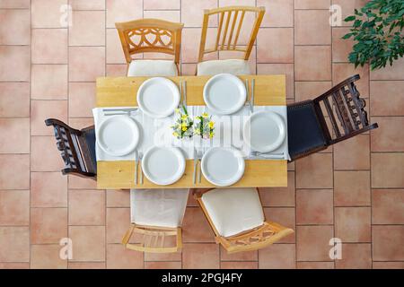 Top view of a place setting with six white plates, cutlery and yellow daffodil flowers on a light wooden table and six different chairs on a terracott Stock Photo