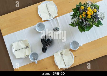 Wooden table with three plates, cups, napkins, coffee pot and a flower bouquet on a white table runner, high angle view from above, selected focus Stock Photo