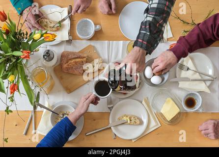 Top view on a group of people sitting on a wooden table and having breakfast together with coffee, bread, eggs, butter and some spring flowers, high a Stock Photo