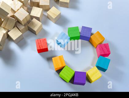 Cubes in different colors together form a circle with a gap, one is red and stands separate, heap of natural wooden cubes in the corner, light blue ba Stock Photo