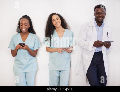 Team of three healthcare professionals lean against wall using digital device Stock Photo