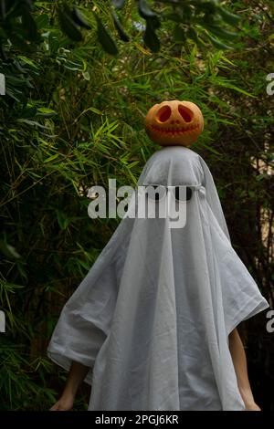 A man dressed in a white ghost costume with sunglasses and a carved pumpkin atop his head for Halloween Stock Photo