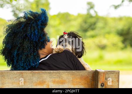 Goiania, Goias, Brazil – March 20, 2023: Photo of a young woman, from the back, sitting on a bench with a shitzu puppy in her lap. Stock Photo