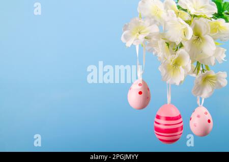 Three delicate pink Easter eggs suspended from a budding branch on a serene blue background, a perfect symbol of spring renewal and Easter holidays Stock Photo