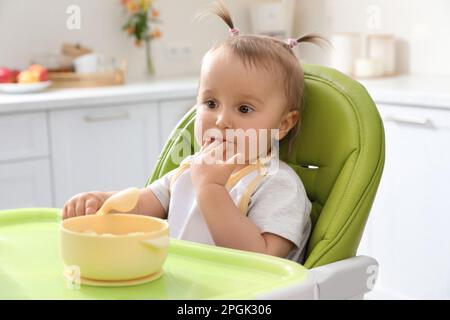 Cute little baby eating food in high chair at kitchen Stock Photo