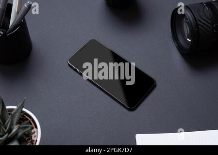 Black office desk table with blank smartphone surounded by camera lens, pens, plant and notepad. Business composition Stock Photo