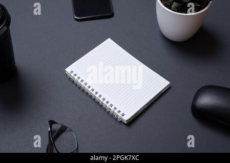 Blank notepad on black work desk witc copy space. Surounded by office supplies, phone, glasses, coffe mug, plant. Low light business composition Stock Photo