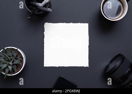 Blank paper frame on black office desk surrounded by phone, camera, coffee mug, plant and pens. Top view, flat lay composition for copy presentation Stock Photo