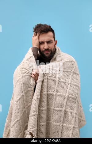 Man wrapped in blanket suffering from headache on light blue background. Cold symptoms Stock Photo