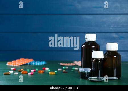 Bottles of cough syrup, dosing spoon, measuring cup and pills on green wooden table. Space for text Stock Photo