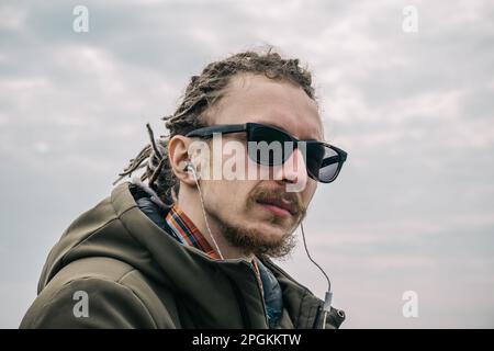 Portrait of cheerful guy with glasses and dreadlocks in-ear headphones or earphones listen to music. Young hipster looking at camera. Stock Photo