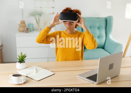 African american young woman wearing using virtual reality metaverse VR glasses headset at home. Girl puts on virtual reality helmet. Technology simulation hi-tech videogame concept Stock Photo