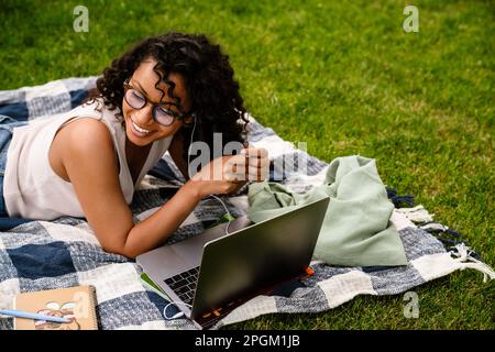 Joyful young african american woman in earphones using laptop while lying on blanket in green park Stock Photo