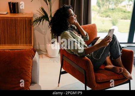 Profile of young smiling african woman in earphones using smartphone while sitting on armchair at home Stock Photo