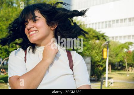 young venezuelan woman university student outdoors, dressed in white with a backpack, shaking her head and smiling. Stock Photo