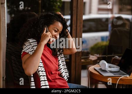 Young black woman smiling and talking on cellphone while sitting at cafe outdoors Stock Photo