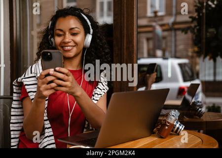 Young black woman using cellphone while working with laptop at cafe outdoors Stock Photo