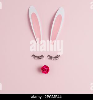 Easter bunny face made of rabbit ears with eyelashes and rose flower on pink background. Happy Easter minimal concept. Stock Photo