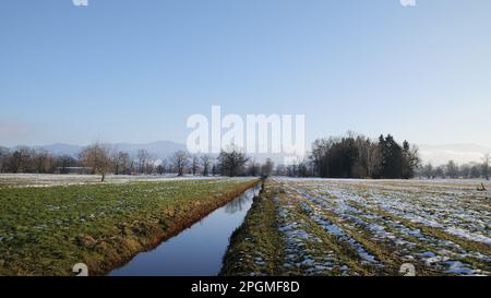 Narrow river in the field of a winter landscape with trees and mountains in the background Stock Photo
