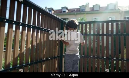 One small boy standing by balcony fence staring outside from second floor with lens flare sunlight. Child holding in wooden bars looking out Stock Photo