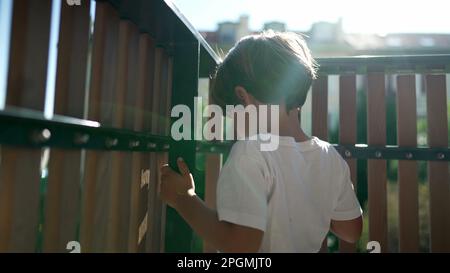 One small boy standing by balcony fence staring outside from second floor with lens flare sunlight. Child holding in wooden bars looking out Stock Photo