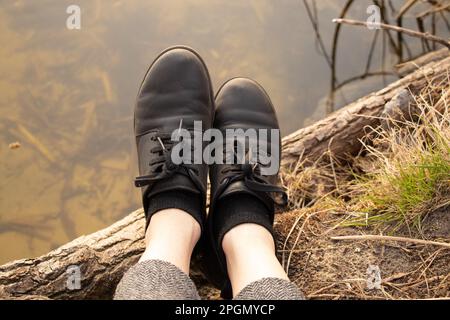 female legs in black leather shoes on the background of the river in spring on vacation in the forest, outdoor recreation Stock Photo