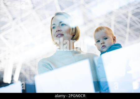 Thoughtful young mother looking trough window holding his infant baby boy child while waiting to board an airplane at airport terminal departure gates Stock Photo