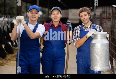 Three dairy farm workers with pitchforks and can of milk stand in cowshed Stock Photo