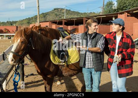 Two skilled man woman stable workers in plaid shirts standing close to saddled horse and discussing upcoming work in countryside club Stock Photo